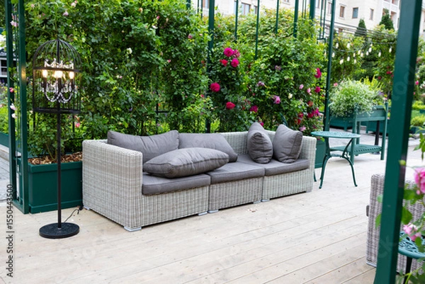 Fototapeta A modern grey sofa with cushions is installed in a glass greenhouse outside. Lush green plants and vibrant flowers surround the living area, creating a serene atmosphere.