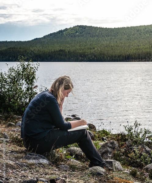 Fototapeta Outdoor woman reading a book while sitting at the shore of a lake on a bright summer day in Sweden. She is enjoying the peace and quiet of nature.