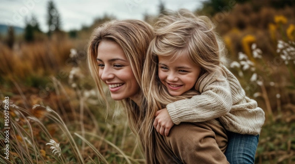 Obraz A child getting a piggyback ride on her mother's back. Both are smiling. They are outdoors. walking in a field. Motherhood. A loving family relationship.
