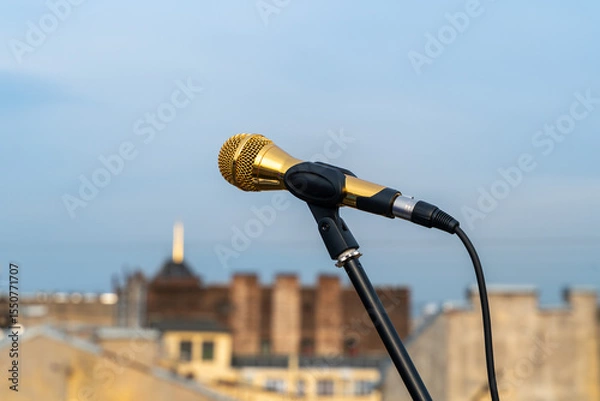 Fototapeta A golden microphone mounted on a stand, set against a backdrop of an urban skyline. the scene combines elements of performance and city life, capturing a moment of potential creativity.
