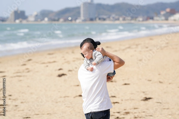 Fototapeta In early March, a Japanese father in his 30s carries his 6-month-old baby on his shoulders while walking along a sandy beach by the sea. A warm springtime scene of family, joy, and connection.