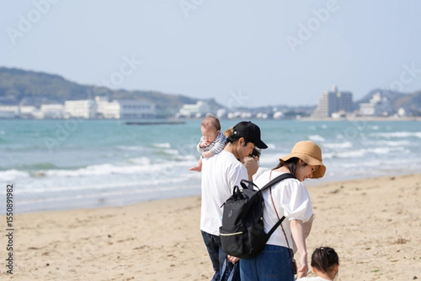 Fototapeta In early March, a 6-month-old baby, a 3-year-old girl, and their Japanese parents in their 30s walk together on a sandy beach by the sea. A springtime moment filled with warmth and connection.
