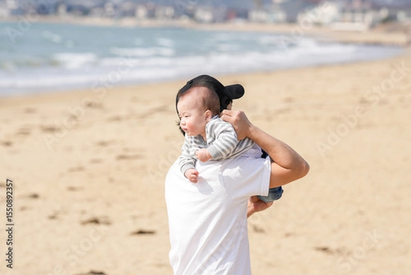 Fototapeta In early March, a Japanese father in his 30s carries his 6-month-old baby on his shoulders while walking along a sandy beach by the sea. A warm springtime scene of family, joy, and connection.