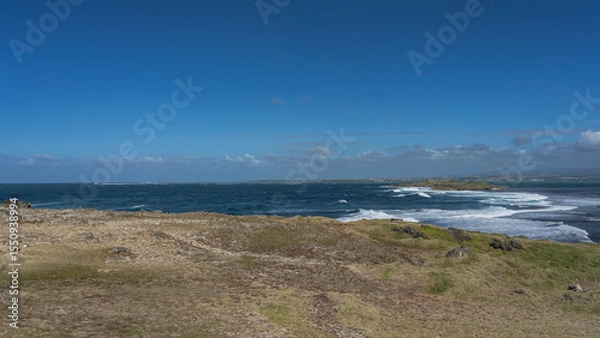 Fototapeta Waves are raging in the blue ocean in the strait between two islets. White foam on the surface of the water. Volcanic boulders, green grass on the shore of the island. Azure sky, clouds. Mauritius.