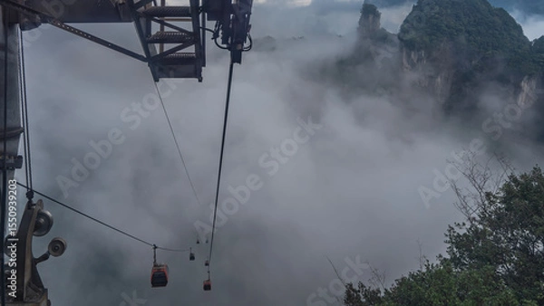 Fototapeta High-altitude cable car. The cabins of the funicular are shrouded in fog. The peaks of the steep mountains above the clouds. Green vegetation and construction details in the foreground. China.
