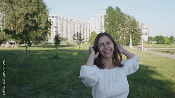 Fototapeta A woman stands in a park, smiling happily while playing with her hair. Modern apartment buildings are visible in the background, highlighting an urban setting.