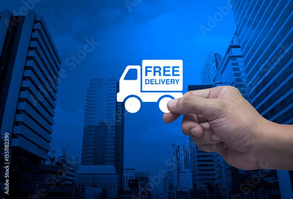 Fototapeta Hand holding free delivery truck flat icon in fingers over modern office city tower and skyscraper, Business transportation service concept