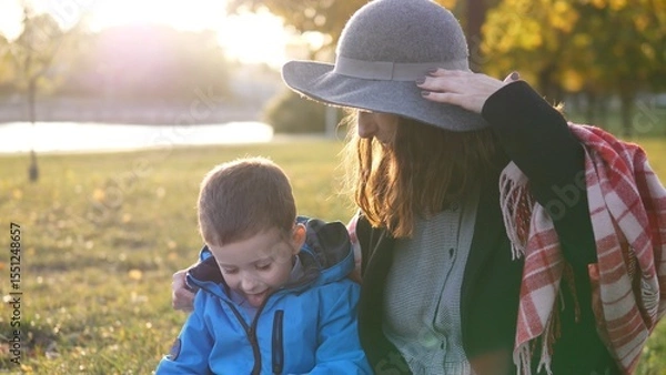 Fototapeta A woman wearing a hat sits beside her young son, both smiling as they share a playful moment on the grass in a park during a sunny autumn afternoon.