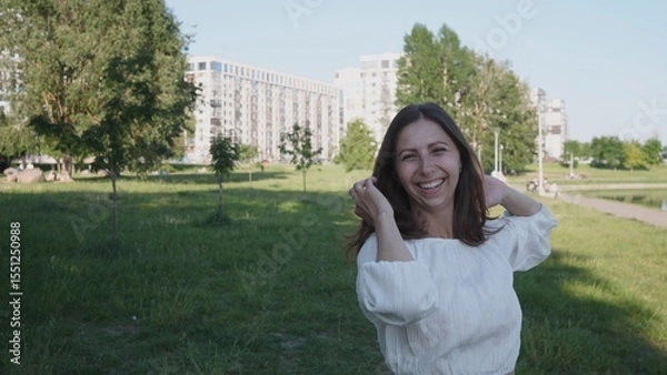 Fototapeta A cheerful woman stands in a lush park, playfully touching her hair while showcasing her smile against a backdrop of urban architecture and greenery.