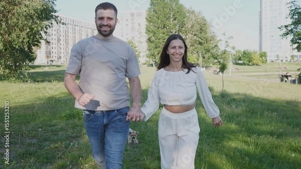 Fototapeta A joyful couple walks hand in hand through a grassy park, smiling and enjoying their time together with a small dog beside them on a sunny day.