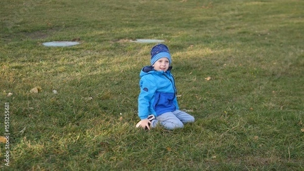 Fototapeta A smiling child sits on the grass, dressed in a blue jacket and hat, enjoying outdoor play in a park during a sunny autumn afternoon.