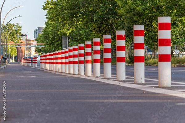 Fototapeta White bollards with red stripes stand in a straight line along a street. This photo shows an urban traffic feature in sunlight.