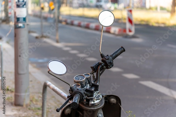 Fototapeta A scooter is parked on the side of the road, the focus is on the handlebars with mirrors. The road and red and white bollards are visible in the background.