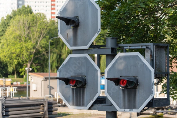 Fototapeta A special waterway traffic light with red signals stands against a green background. The technical device appears industrial and unusual in the context of the cityscape