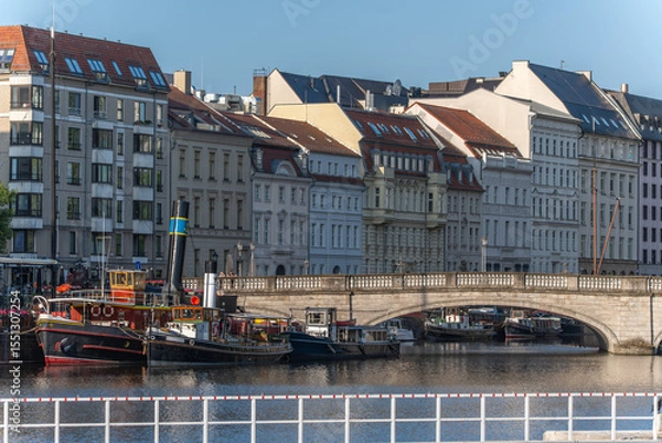 Fototapeta Historic buildings and houseboats line the riverbank in front of an old stone bridge in Berlin. An idyllic scene of urban waterways in sunlight.