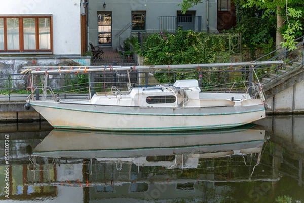 Fototapeta A white sailboat rests peacefully in the water in front of a residential building. The reflection in the water lends the image a tranquil atmosphere.