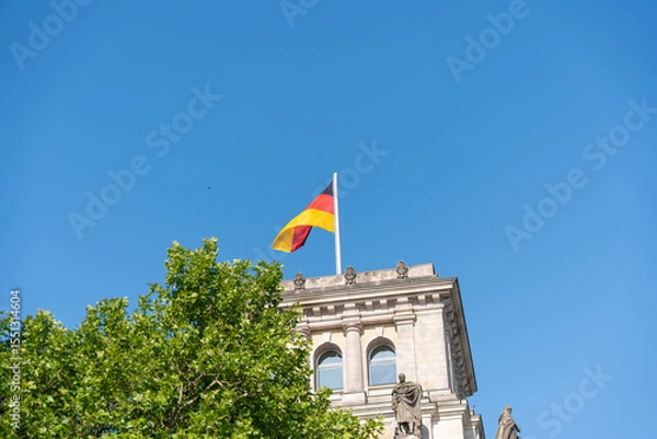 Fototapeta The German national flag waves atop a historic building with statues and columns. Bright blue sky and green tree leaves frame the scene.