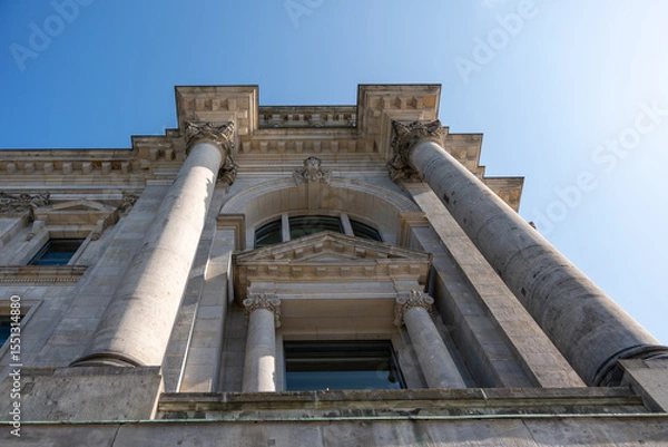 Fototapeta Neoclassical Facade with Columns. Upward view of a neoclassical building with large stone columns and ornate details. The clear blue sky frames the monumental structure.