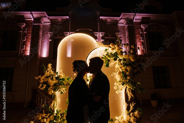 Obraz Silhouettes of bride and groom in front of a wedding arch decorated with flowers