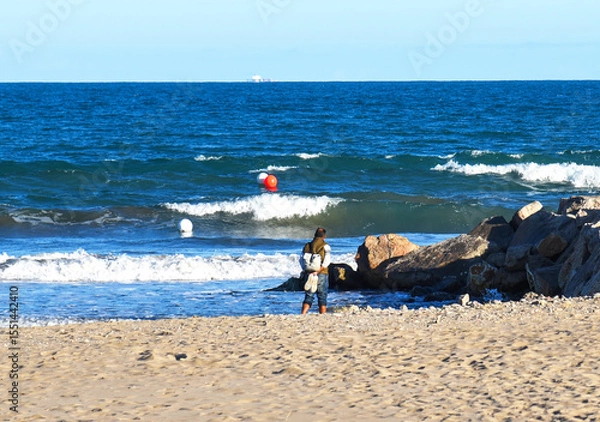 Obraz Man at sea beach on blu sky. Back side of European man 40-70s in jacket, standing, waiting or thinking about something in feeling lonely sadness. Male at beach, man standing in sea, waves on his feet.