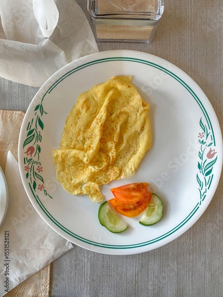 Fototapeta Close up top down view of plain omelette served with tomato and cucumber slices on floral rim white plate. Minimalist breakfast at hotel guesthouse casa particular, Cienfuegos, Cuba. Natural lighting
