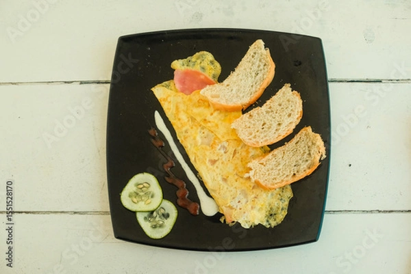 Fototapeta Simple breakfast of omelette, sliced bread, cucumber on black plate on rustic white wooden table. Morning meal in local restaurant, close up top-down view. Travel food photography Varadero, Cuba
