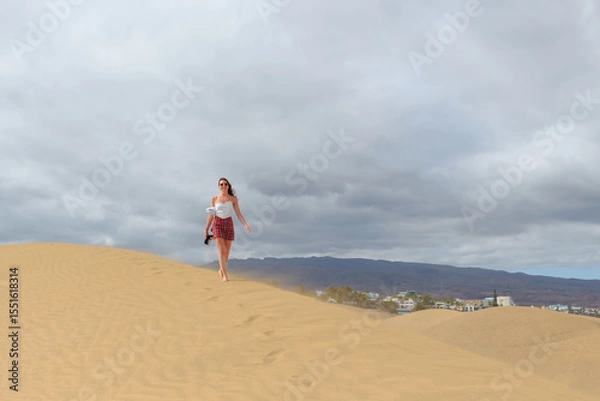 Fototapeta A woman in a white top and plaid skirt walks gracefully atop a sand dune, with distant mountains and buildings under cloudy skies.
