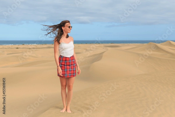 Fototapeta A young woman in a plaid skirt and top enjoys the warm breeze while standing barefoot in a sandy desert, with ocean waves visible in the background.