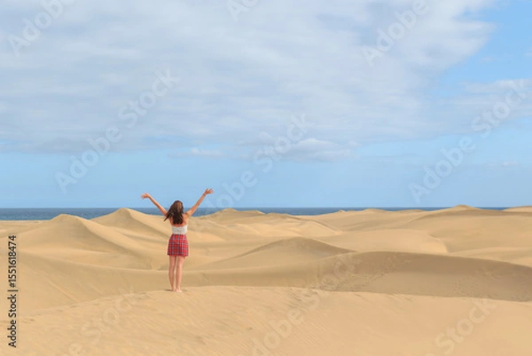 Fototapeta A person stands barefoot on golden sand dunes, arms raised in celebration, with the ocean visible in the background under a partly cloudy sky.