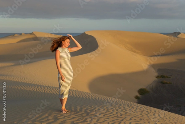 Fototapeta A woman strolls barefoot across golden sand dunes, enjoying the warm glow of the setting sun and gentle breeze in a tranquil desert landscape.