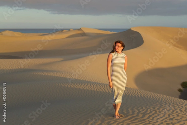 Fototapeta A woman strolls elegantly among golden sand dunes as the sun sets, casting long shadows across the landscape by the ocean.