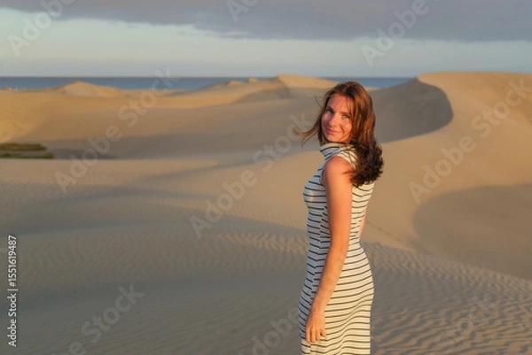 Fototapeta A woman in a striped dress walks gracefully on the sandy dunes as the sun sets, creating a serene ambiance in the coastal landscape.