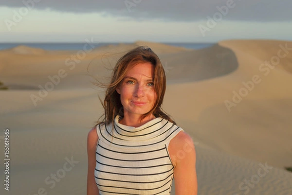 Fototapeta A woman with long hair poses in desert sand dunes as the sun sets, creating a tranquil and scenic atmosphere with a beach in the background.