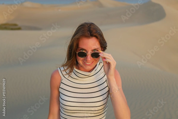 Fototapeta A woman with sunglasses smiles while standing on golden sand dunes during a warm afternoon in a remote desert landscape.