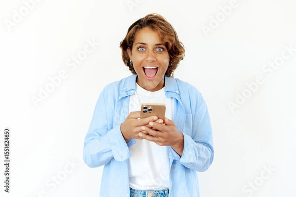 Obraz Shocked blue-eyed brazilian young adult woman enthusiastically using smartphone, reacting with big smile and excitement, standing on isolated white background and amazed looking at the camera