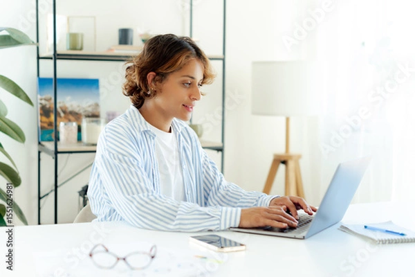 Obraz Pretty brazilian creative professional typing on a laptop while working in her modern office. Young female freelancer with glasses focused on digital project or communication in home office setup
