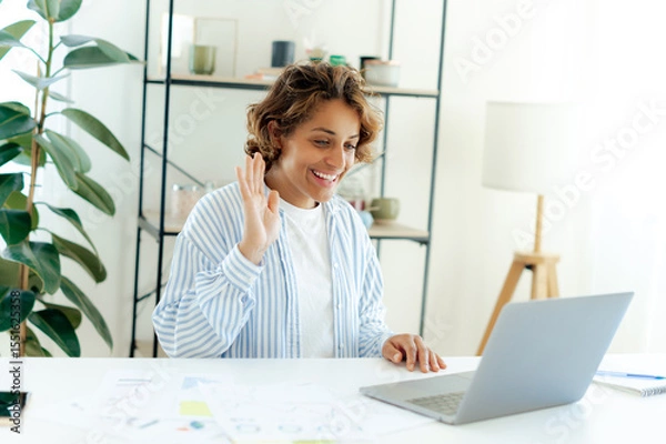 Obraz Positive brazilian woman waving at laptop screen during video call while working remotely in a modern space. Successful businesswoman attending online meeting or virtual conference, smiles