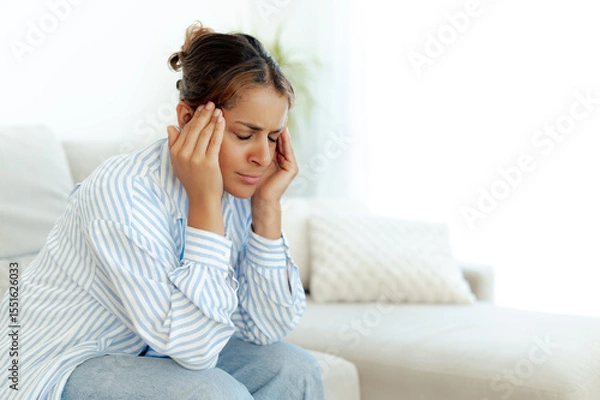 Obraz Upset brazilian woman with eyes closed and hands on temples, suffering from a strong headache or stress at home. Dressed in a casual clothes, she sits on couch feeling discomfort