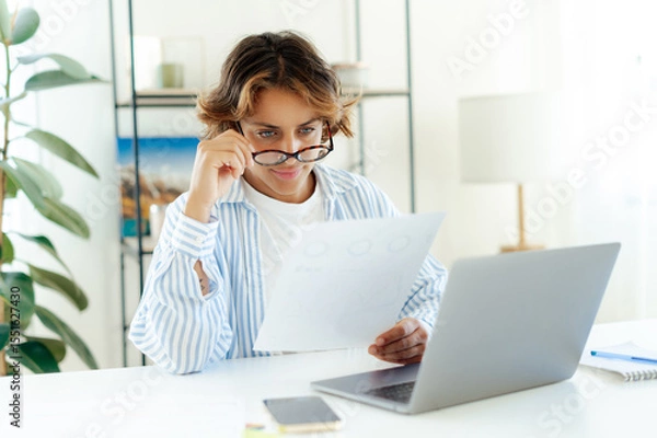 Obraz Serious brazilian successful businesswoman with glassed reading printed report while working on laptop at home. Focused professional analyzing data or financial charts during remote work in apartment