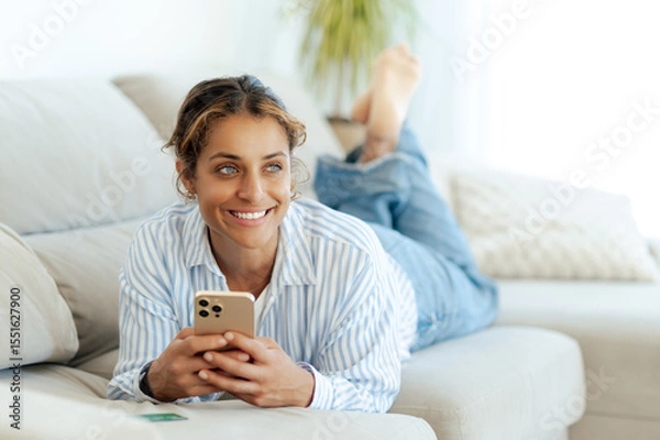 Obraz Pretty brazilian blue-eyed woman in casual wear is lying on couch, holding smartphone and smiling. She is relaxed and happy while browsing or chatting online on a sofa ina living room