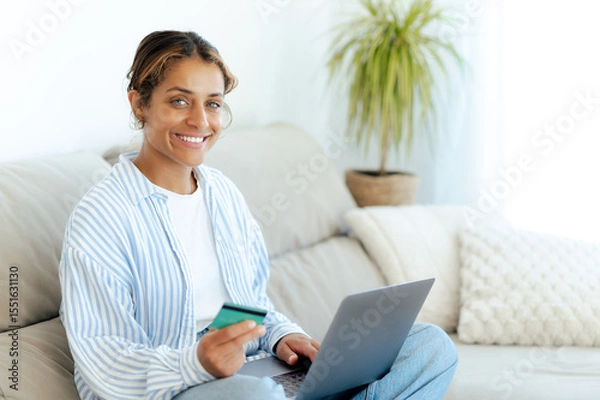 Obraz Positive brazilian woman in casual clothes sitting on couch at home, smiling while holding a credit card and using a laptop. She enjoys online shopping in a relaxed, cozy setting in a living room