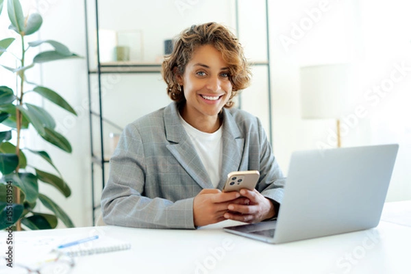 Obraz Smiling brazilian female company employee holding smartphone while working at a workplace on a laptop. Confident young businesswoman managing tasks and communication in modern office using her phone