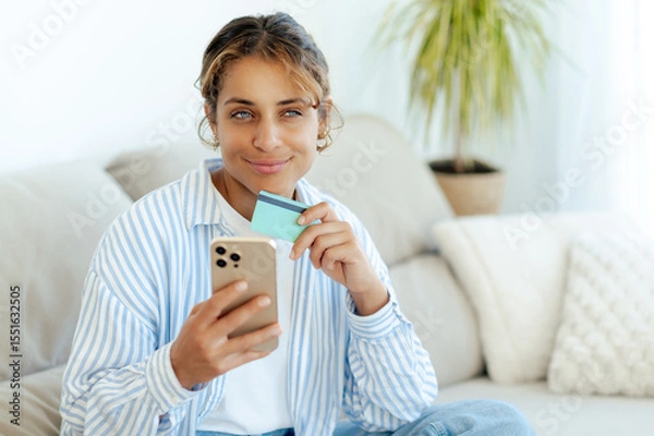 Obraz Positive brazilian woman holding credit card and smartphone, making online purchase or banking from home sitting at sofa. She looks pleased and thoughtful while shopping or managing finances on mobile