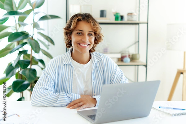 Obraz Beautiful brazilian professional woman smiling confidently while sitting at desk with laptop during online work. Friendly freelancer or online coach posing at a workplace, looking and smiles at camera