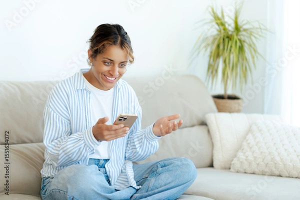 Obraz Excited happy brazilian woman sits cross-legged on sofa, laughing and chatting during video call on smartphone. Relaxed she enjoys a spending time scrolling social media and reads good news at home