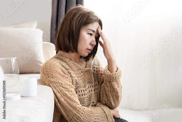 Fototapeta Stressed Asian woman feeling worried about chronic migraine, holding her forehead in pain, showing signs of ongoing headache and discomfort affecting her daily life