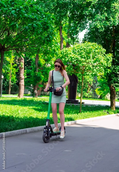 Obraz Eco-friendly transport concept. A female enjoys riding a shared electric kick scooter in park.Summer city leisure, sustainable lifestyle in an urban environment.