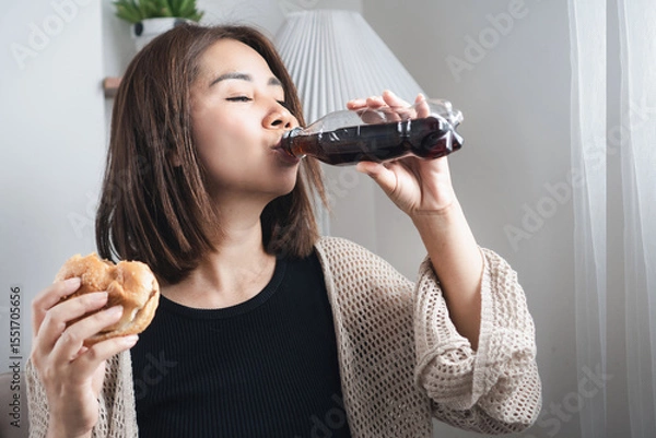 Obraz Asian woman drinking soda and holding a burger in both hands, showing emotional eating and unhealthy food choices high in sugar, salt, and fat