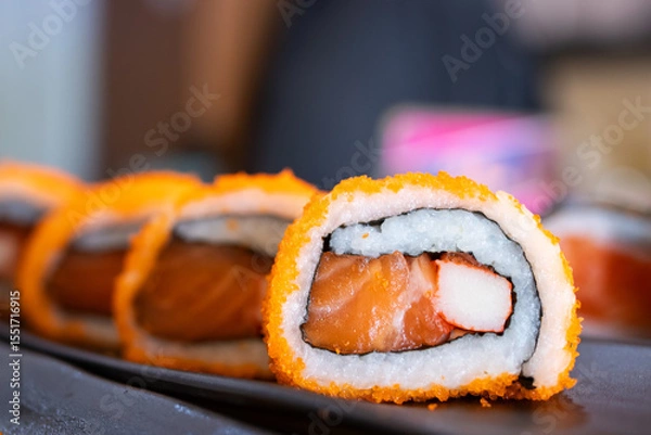 Fototapeta Sushi roll with salmon, crab stick, and rice, coated with orange tobiko, served on a black tray in a Japanese restaurant setting.