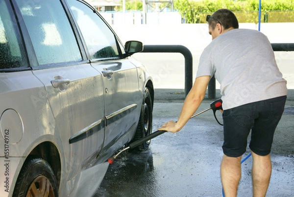 Fototapeta A young man bends over and washes the underside of a silver-gray passenger car with a high-pressure water gun at a self-service car wash in an open-type bay. Self-cleaning of personal transport